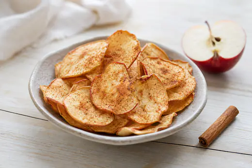 Tunna krispiga äppelchips med kanel i en djup skål, med ett halvt rött äpple bredvid