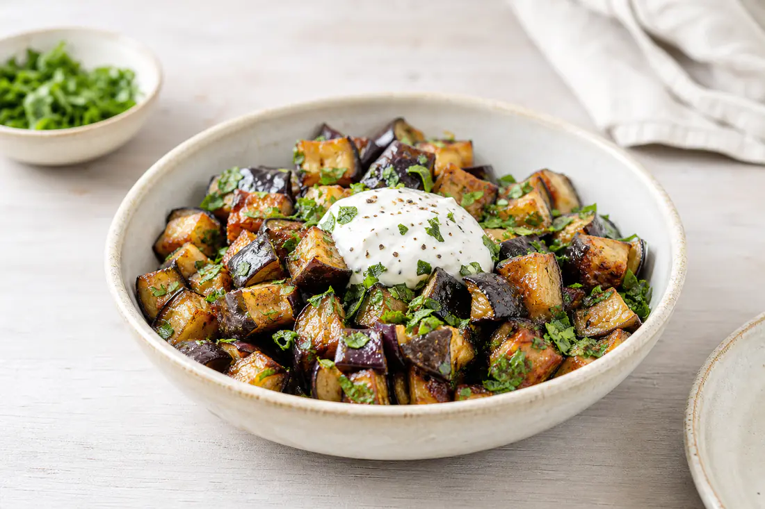 Rostade auberginetärningar med gyllene kanter i en skål, toppade med persilja och en klick yoghurtsås