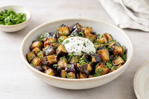 Rostade auberginetärningar med gyllene kanter i en skål, toppade med persilja och en klick yoghurtsås