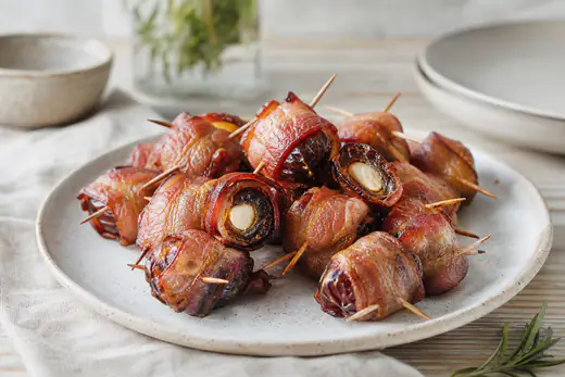 Baconlindade dadlar med tandpetare på ett serveringsfat, några uppskurna så att mandel syns inuti