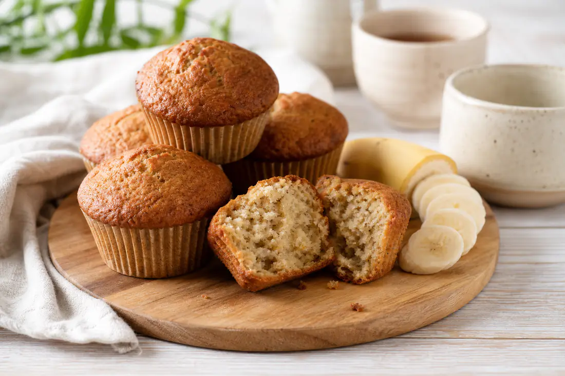 Gyllenbruna bananmuffins i pappersformar på ett träbräde, en muffins delad på mitten och skivad banan bredvid