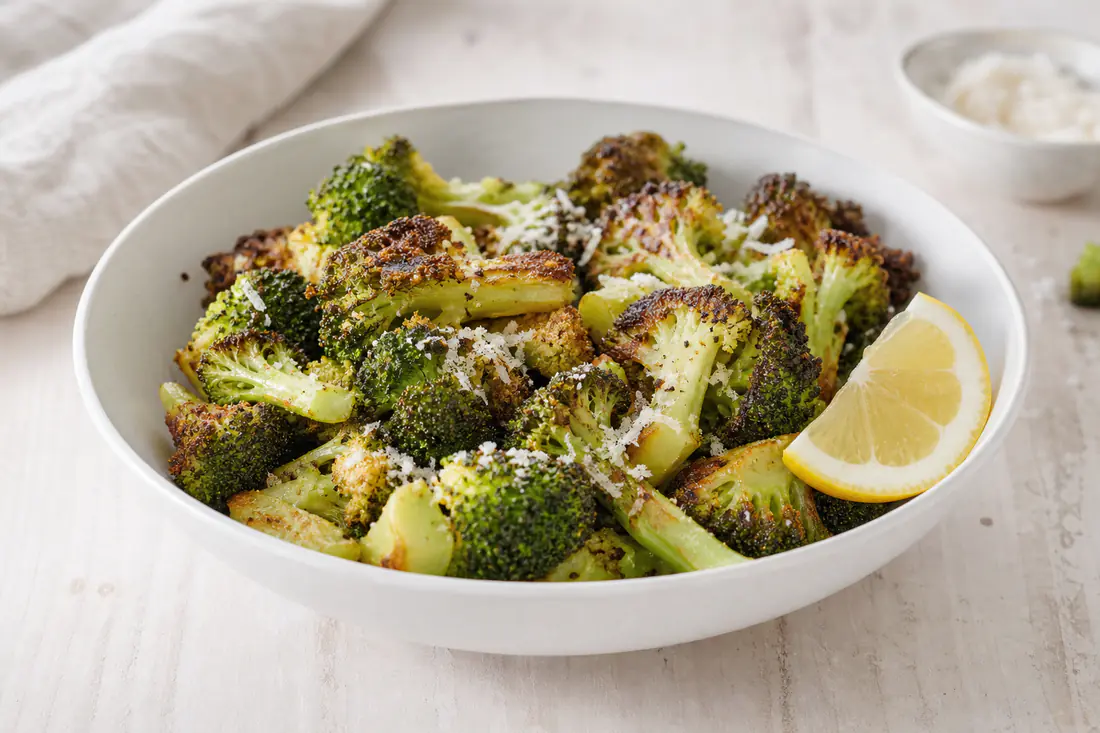 Rostade broccolibuketter med brynta kanter och riven parmesan i en vit skål med citronklyfta