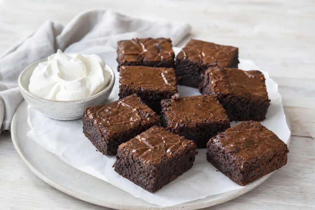 Brownierutor med blank, sprucken yta och kladdig mitt på ett bakplåtspapper bredvid en skål med vispad grädde