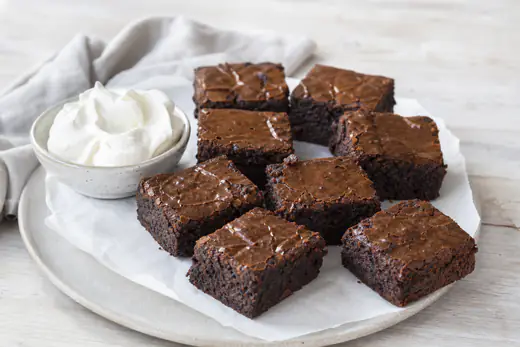 Brownierutor med blank, sprucken yta och kladdig mitt på ett bakplåtspapper bredvid en skål med vispad grädde