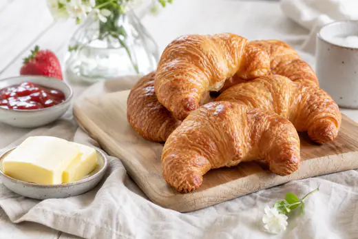 Gyllenbruna croissanter av smördeg på ett träbräde med smör och en skål jordgubbssylt