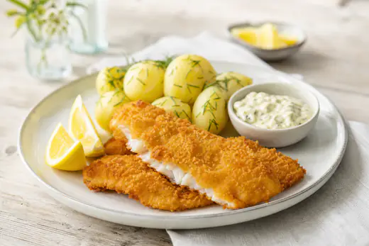 Gyllene panerade fiskfiléer på en tallrik med citronklyftor, kokt potatis och remouladsås