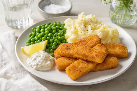 Gyllene fiskpinnar på en tallrik med potatismos, gröna ärtor, remouladsås och citronklyfta
