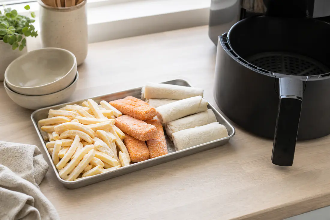 Frysta pommes, fiskpinnar och vårrullar med frost på en bricka bredvid en airfryerkorg