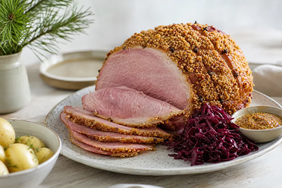 Griljerad julskinka med gyllene ströbrödsyta, delvis skivad på ett fat med senap och rödkål