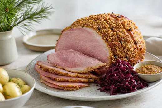 Griljerad julskinka med gyllene ströbrödsyta, delvis skivad på ett fat med senap och rödkål