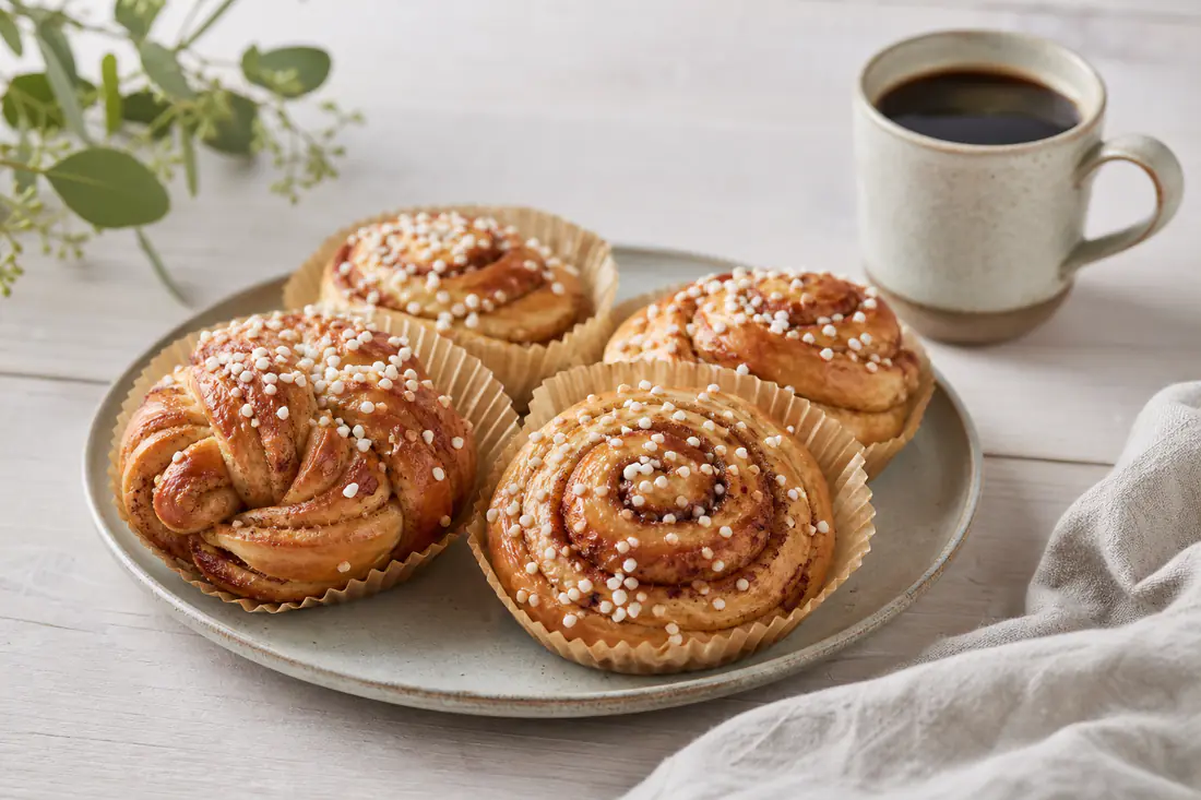 Nybakade kanelbullar med pärlsocker i pappersformar på ett fat bredvid en kopp kaffe