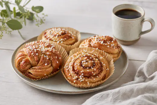 Nybakade kanelbullar med pärlsocker i pappersformar på ett fat bredvid en kopp kaffe
