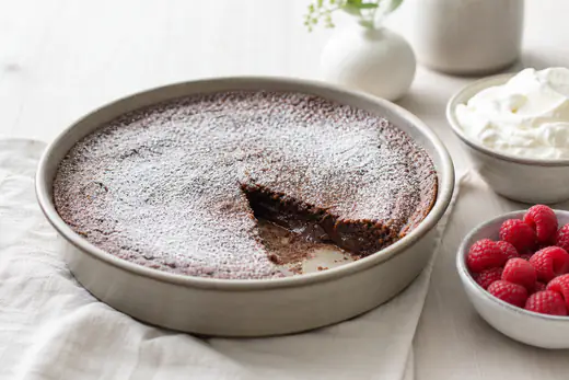Kladdkaka med florsocker i en rund form där en bit är uppskuren och visar kladdig mitt, med vispad grädde och hallon bredvid