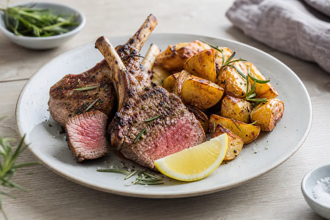 Stekta lammkotletter med rosa insida och färsk rosmarin på en tallrik med rostad potatis och en citronklyfta