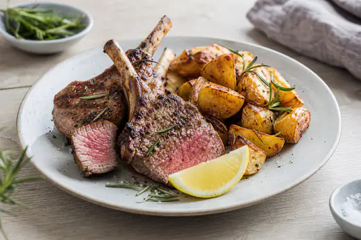 Stekta lammkotletter med rosa insida och färsk rosmarin på en tallrik med rostad potatis och en citronklyfta