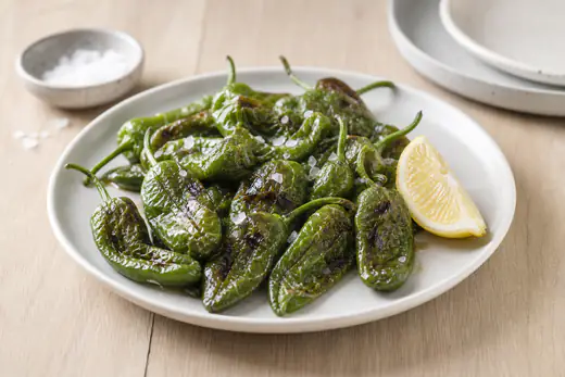 Små gröna padrones med blåsor och mörka fläckar, saltade med flingsalt på en liten tallrik med citronklyfta