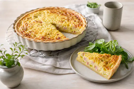 Gyllene skinkpaj med ost och purjolök i en liten rund pajform, en bit uppskuren bredvid en grönsallad