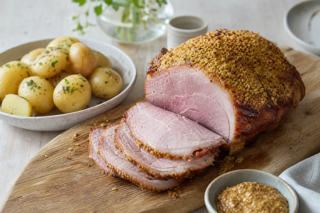Glaserad rimmad picnicbog med gyllene senapsskorpa, delvis skivad på en skärbräda med kokt potatis och senap