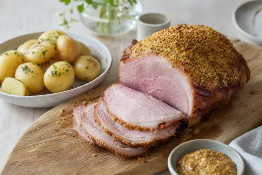 Glaserad rimmad picnicbog med gyllene senapsskorpa, delvis skivad på en skärbräda med kokt potatis och senap