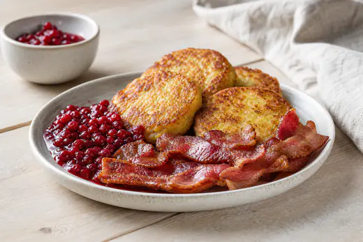 Gyllene potatisbullar på en tallrik med rårörda lingon och stekt bacon