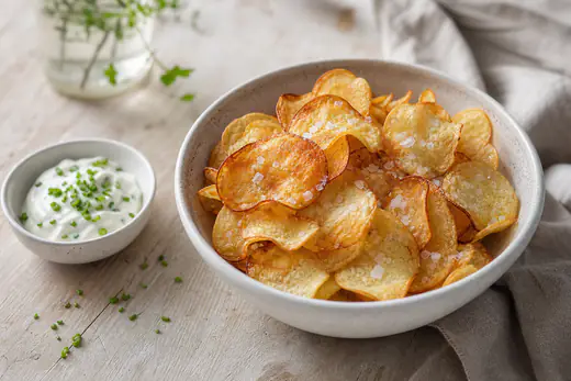 Hemgjorda tunna potatischips, gyllene och krispiga, i en skål med flingsalt och en liten skål dipp