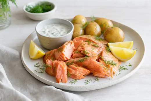 Varmrökt lax i bitar med dill och citronklyftor på en tallrik med kokt färskpotatis och kall sås