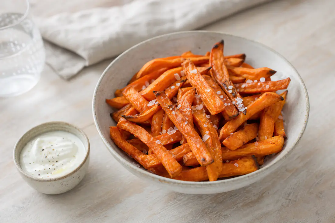 Orange sötpotatisfrites med flingsalt i en skål med en dippsås av yoghurt bredvid