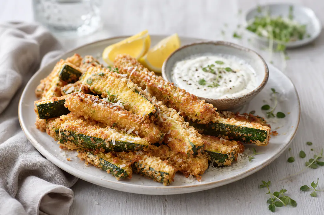 Gyllene panerade squashstavar med panko och parmesan på ett fat med en skål vitlöksdipp och citronklyftor