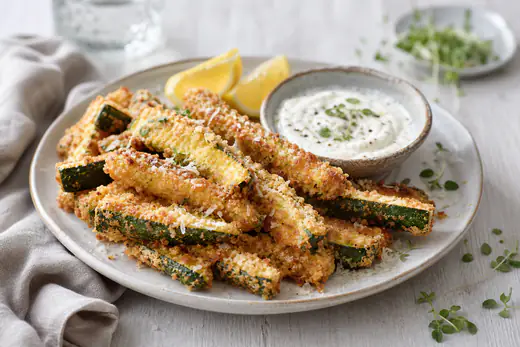 Gyllene panerade squashstavar med panko och parmesan på ett fat med en skål vitlöksdipp och citronklyftor
