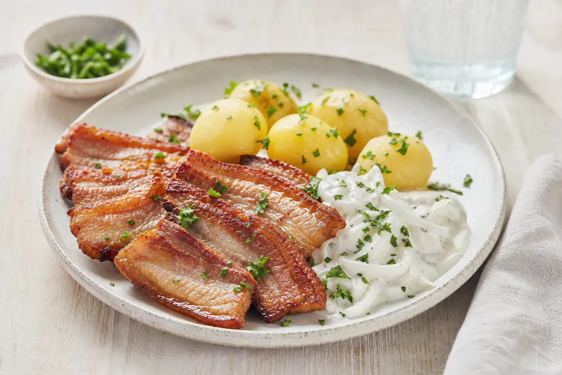 Krispiga skivor stekt fläsk på en tallrik med kokt potatis, vit löksås och persilja