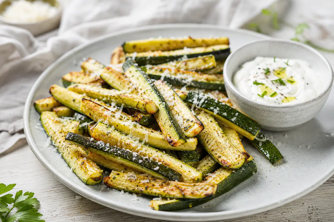 Rostade zucchinistavar med gyllene kanter och riven parmesan på ett fat med en skål yoghurtdipp