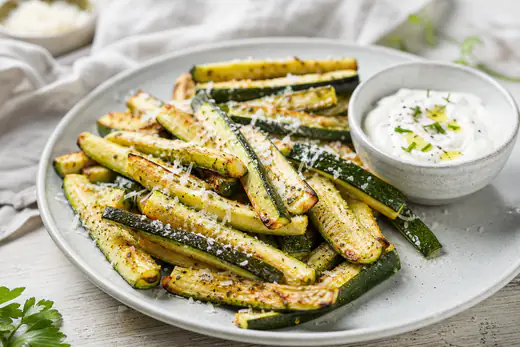 Rostade zucchinistavar med gyllene kanter och riven parmesan på ett fat med en skål yoghurtdipp